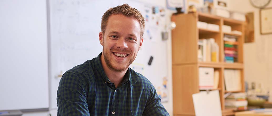 Happy bearded male teacher at desk looking to camera